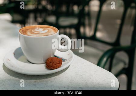 Cappuccino cremoso in una tazza bianca servito con un biscotto su un tavolo luminoso del caffè. Atmosfera calda di pausa caffè e concetto di stile di vita mattutino accogliente. Foto Stock