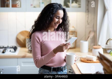 Comunicazione mobile. Giovane bella donna con smartphone e bere caffè in cucina Foto Stock