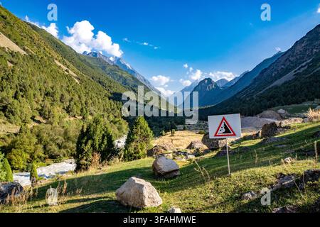 Un cartello stradale di avvertimento con un simbolo di frana si trova accanto a un sentiero di montagna che si affaccia su una verde valle alpina, attraversata da un fiume dalle acque veloci Foto Stock