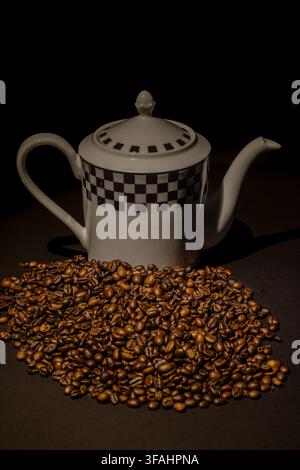 Caffettiera in ceramica bianca a motivi che si trova dietro un grande mucchio di chicchi di caffè tostati su uno sfondo scuro, illuminata da una luce calda e drammatica. Foto Stock