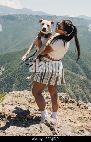 Donna che abbraccia un cane Pitbull e gode della vista sul paesaggio montano in una giornata di sole Foto Stock