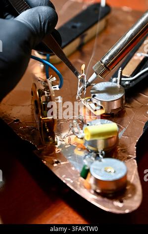 Fotografia pubblicitaria in primo piano di un tecnico che indossa guanti protettivi neri, utilizza una pinzetta per tenere un componente durante la saldatura del cablaggio di una chitarra elettrica su un pickguard schermato in rame. Un sottile filo di fumo si alza dalla punta del saldatore mentre tocca il contatto del potenziometro, enfatizzando la precisione artigianale e il lavoro di riparazione. Foto Stock
