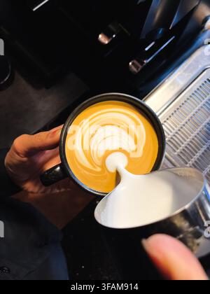 Scatto dall'alto di un barista che versa latte vaporizzato schiumoso per creare una delicata latte art in una tazza nera. Preparazione professionale del caffè. Foto Stock