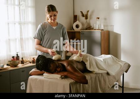 Massaggiatrice che massaggia una donna nera con un impasto alle erbe in un centro benessere Foto Stock