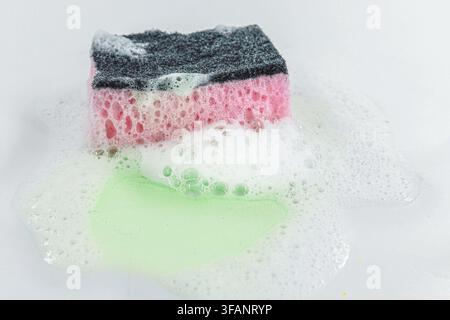 Spugna da cucina rosa usata coperta di schiuma di sapone su una superficie bianca bagnata Foto Stock