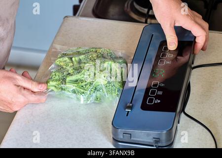 Macchina per la sigillatura sottovuoto domestica che sigilla broccoli freschi in un sacchetto di plastica, mostrando il processo di conservazione degli alimenti e lo stoccaggio in cucina sul piano di lavoro. Foto Stock