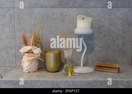 Scaffale da bagno accogliente con diffusore di bacche profumato, candela, spazzola naturale per il corpo e decorazione con fiori essiccati su piastrelle grigie. Concetto di spa domestica, relax Foto Stock