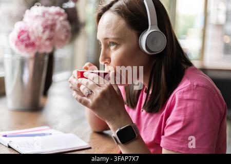 Donna con cuffie che beve caffè da una tazza rossa in un caffè Foto Stock