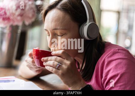 Donna che beve caffè da una tazza rossa mentre indossa le cuffie Foto Stock