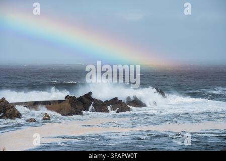 Arcobaleno che si inarca sopra le onde tempestose dell'oceano che si infrangono sugli scogli Foto Stock