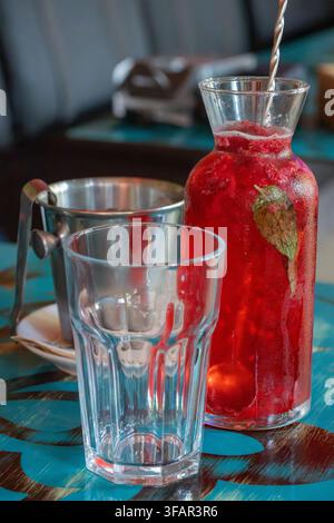 Una bevanda rinfrescante ai frutti di bosco rossi in una caraffa di vetro è presentata insieme a un bicchiere sfaccettato vuoto e un secchiello di ghiaccio in metallo su un tavolo da caffè blu a motivi Foto Stock