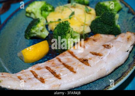 un filetto di pesce bianco grigliato sano con segni di griglia distinti, servito su un piatto di ceramica blu insieme a cimette di broccoli al vapore, purè di patate cremoso p Foto Stock