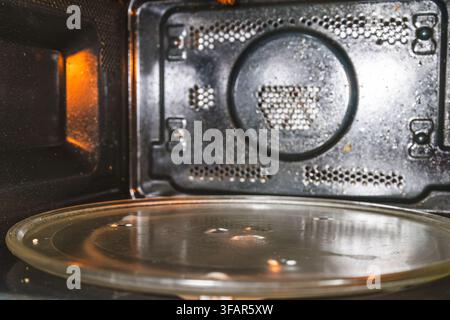 Un dettagliato scatto interno di un forno a microonde che mostra macchie di grasso, schizzi di sporco e un vassoio rotante in vetro all'interno di una moderna cucina domestica Foto Stock