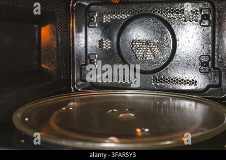 Un primo piano interno di un forno a microonde sporco che mostra macchie di grasso scure, schizzi di cibo e un piatto girevole in vetro Foto Stock