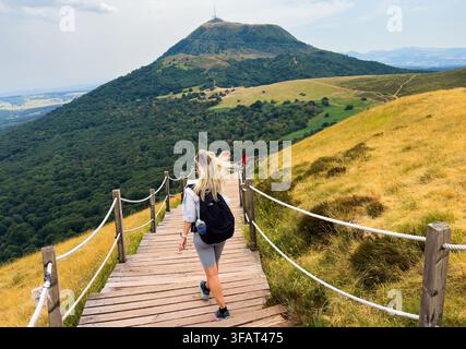 donna che fa escursione su passerella di legno in una foresta verde con ringhiere di corda, prospettiva in prima persona da dietro di lei, atmosfera avventurosa, ambientazione di sentiero di montagna Foto Stock