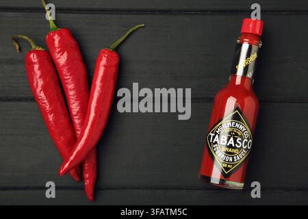 Mykolaiv, Ucraina - 19 agosto 2026: Una bottiglia di salsa al pepe Tabasco con peperoncino rosso su sfondo di legno nero. Vista dall'alto Foto Stock