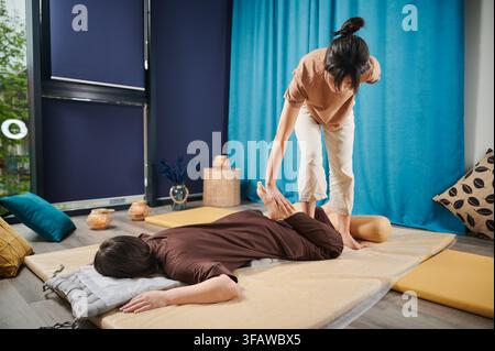 Massaggiatrice che fa un massaggio yoga tailandese. Terapeuta donna che fa un massaggio tradizionale, allungando il corpo di una paziente. I clienti sono sdraiati sul tappetino e si godono un massaggio alle mani in un'atmosfera tranquilla. Foto Stock