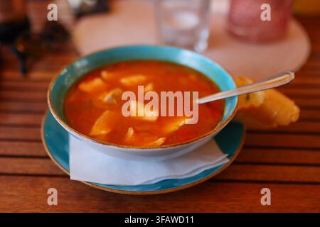 Zuppa di pesce tradizionale con verdure servita in una ciotola di ceramica con pane su un tavolo di ristorante in legno. Foto Stock