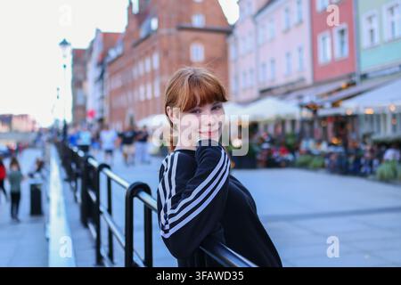 Giovane donna sorridente in posa su una passeggiata sul lungomare con edifici storici colorati e ristoranti sullo sfondo. Foto Stock