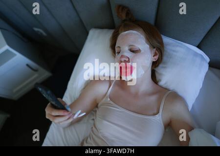 Giovane donna che si rilassa a letto con una maschera in foglio e un patch per le labbra mentre utilizza lo smartphone durante la sua routine di cura della pelle a casa. Foto Stock