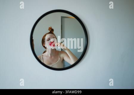 Giovane donna che applica una maschera viso in foglio e un patch per le labbra mentre si guarda allo specchio durante la sua routine di cura della pelle a casa. Foto Stock