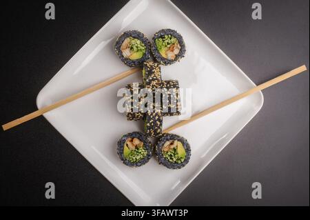 Rotoli di sushi al sesamo nero che formano un motivo creativo su un piatto quadrato bianco con bacchette di legno, scatto ravvicinato. Foto Stock