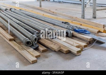 Tubi e raccordi di ponteggio in alluminio smontati sono disposti su assi di legno pallido su un pavimento polveroso, accanto a una cazzuola per intonaco blu e una sega a mano. Th Foto Stock