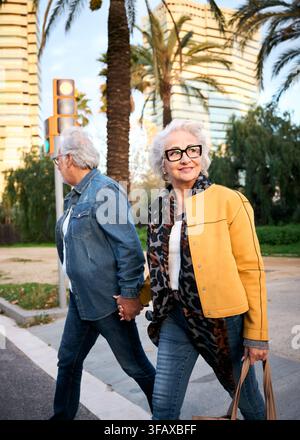Coppia di anziani che si tengono per mano mentre attraversano una strada moderna della città Foto Stock