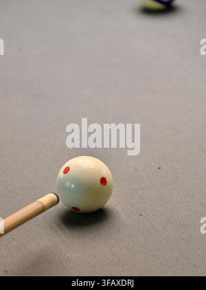 Una fotografia in primo piano verticale di una stecca da biliardo posizionata per colpire una palla bianca segnata con punti rossi su una superficie moderna di tavolo da biliardo in feltro grigio. Foto Stock
