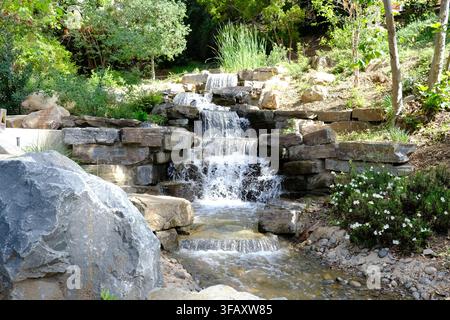 Una piccola cascata paesaggistica scende a cascata su rocce attraverso un lussureggiante giardino. Foto Stock
