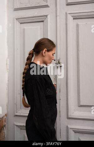 Vista di profilo di una giovane donna con i capelli intrecciati in piedi accanto a una porta grigia alla luce del giorno Foto Stock