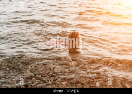 Donna, acqua, nuoto. Giovane donna godersi un bagno rinfrescante in un oceano tranquillo con fondo di ciottoli sotto una luce dorata. Foto Stock
