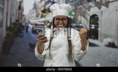 Donna chef con camice bianco e toque, sorride mentre tiene uno smartphone e fa un gesto di pugno pompante su una strada con edifici e auto sfocate; Foto Stock