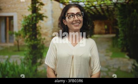 Donna con occhiali che sorride alla telecamera nel cortile di un edificio con camicetta bianca visibile e posa rilassata incorniciata da una pergola; riflessione di contentezza. Foto Stock