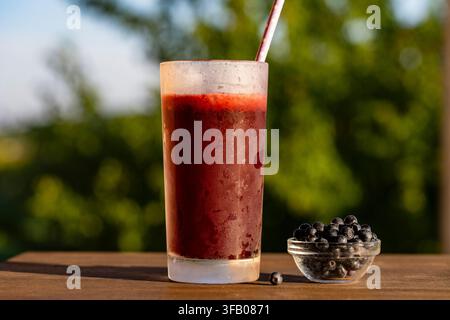 Frullato di mirtilli fresco in vetro, all'aperto, primo piano. Frullato o succo di frutti rossi raffreddato in vetro smerigliato con cannuccia di carta accanto a una piccola ciotola di frutti freschi Foto Stock