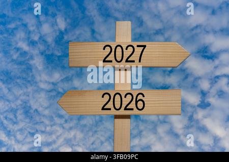 Segnale stradale in legno con gli anni 2026 e 2027 che indicano direzioni opposte. Concetto di transizione temporale dall'anno 2026 all'anno 2027 su un indicatore stradale in legno con c Foto Stock
