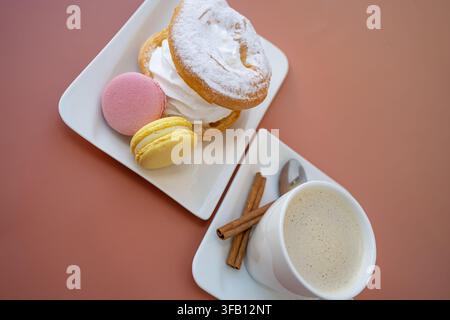 Prospettiva dall'alto di un set per colazione dolce che include una tazza di caffè, macarons colorati, un biscotto di meringa soffice e bastoncini di cannella rustici Foto Stock