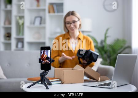Giovane donna che crea contenuti per il suo canale di social media, sblocca un nuovo visore di realtà virtuale e registra un video sul suo smartphone con un laptop nelle vicinanze Foto Stock