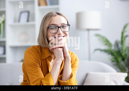 Donna bionda con taglio a caschetto e occhiali che sorride con sicurezza su un comodo divano in un ufficio domestico moderno e luminoso, abbigliamento smart-casual e rilassata durante il lavoro a distanza o una videochiamata Foto Stock
