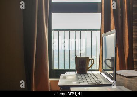 Una confortevole postazione da ufficio domestica con un laptop aperto e una tazza di caffè posizionati su un tavolo accanto a una grande finestra luminosa con tende marroni. Foto Stock