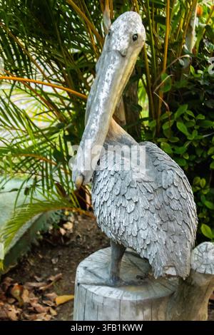 Statua da giardino di un pellicano grigio consumata dalle intemperie, in piedi su un ceppo d’albero tra lussureggianti foglie di palme tropicali e piante verdi all’aperto Foto Stock