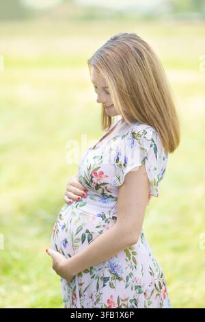 Ritratto di profilo di una donna incinta sorridente in un vestito estivo a stampa floreale che tiene il pancione e guarda amorevolmente verso il basso in un ambiente park all'aperto Foto Stock