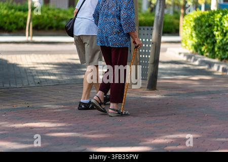Uomo e donna anziani che camminano insieme su un marciapiede pavimentato ombreggiato mentre la donna usa un bastone Foto Stock