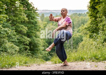 Uomo maturo pratica tai chi su un sentiero boschivo, bilanciandosi con calma in una posa di movimento consapevole che supporta il benessere, la salute olistica, l'invecchiamento sano, un Foto Stock