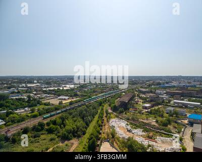 Un colpo aereo con droni ad ampio angolo cattura un lungo treno merci sui binari ferroviari circondato da alberi verdi, edifici industriali, strutture vecchie e un vasto cielo cittadino Foto Stock