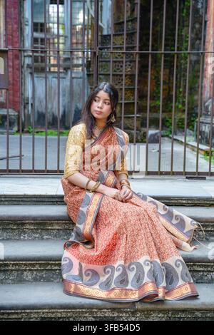 Una persona è seduta sui gradini all'aperto indossando un tradizionale saree con disegni intricati e una camicetta giallo senape. Foto Stock