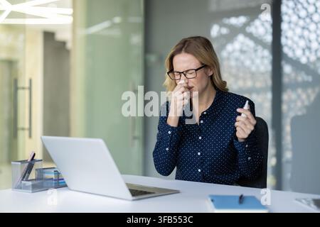 Donna d'affari con occhiali che sperimenta sintomi di malattia, tiene un fazzoletto al naso e uno spray nasale, seduta a una scrivania con un laptop in un ufficio moderno, alle prese con la malattia Foto Stock
