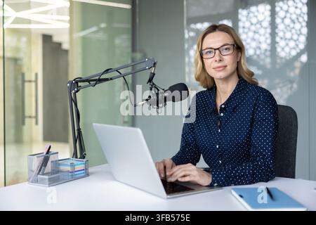 Donna che registra un episodio di podcast in un ufficio moderno, seduta a una scrivania con laptop e microfono professionale, concentrata sulla creazione di contenuti audio digitali per lo streaming e la pubblicazione Foto Stock