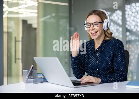 Giovane donna professionista con occhiali e cuffie che sorride e saluta durante una videoconferenza su laptop in un moderno ufficio di coworking, rappresentando il telelavoro e la collaborazione virtuali Foto Stock