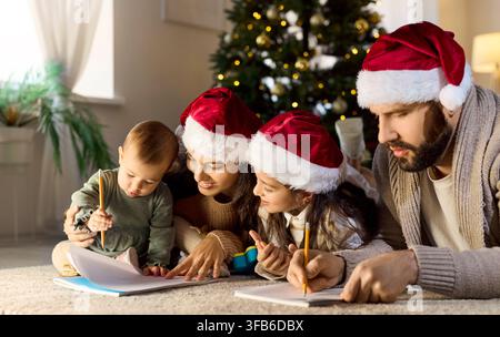 Famiglia felice che disegna sul pavimento di casa a Babbo Natale nei cappelli vicino al luccicante albero di Natale Foto Stock